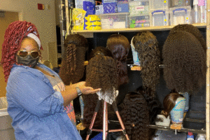 Destinee Steele, a Black woman with red hair in blue jean shirt with wigs on styling heads.