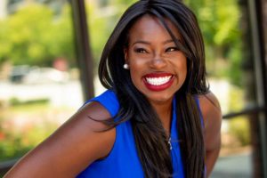 African American woman with long black hair smiling in blue sleeveless shirt outside on sunny day