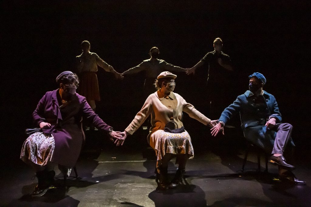 Students wearing masks hold hands while seated onstage.