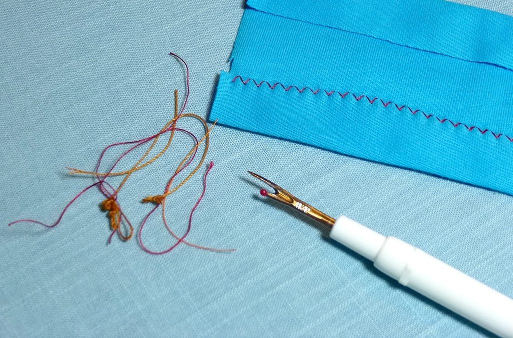 A seam ripper has one long side with a pointed end and one short side with a red ballpoint.